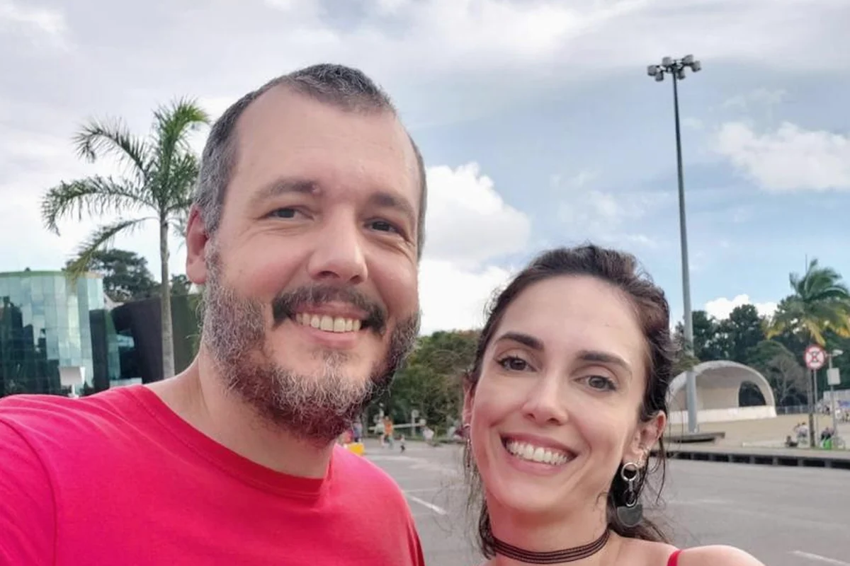 Ricardo e Isabele lado a lado ao ar livre no fim da tarde, os dois sorrindo para a câmera, com céu nublado, palmeiras e um prédio de vidro ao fundo.